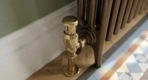 A close-up shot of a polished brass thermostatic radiator valve with numbered heat settings attached to a dark bronze column radiator.