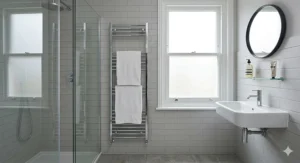 A modern polished chrome vertical heated towel rail, holding two neat white towels, is mounted on light grey subway tiles in a compact en-suite bathroom.