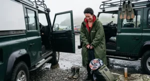 A wild swimmer using a spacious insulated changing robe to get changed privately in a coastal car park next to green off-road vehicles.