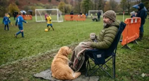 A spectator using a portable heated chair while watching a grassroots football match in autumn.